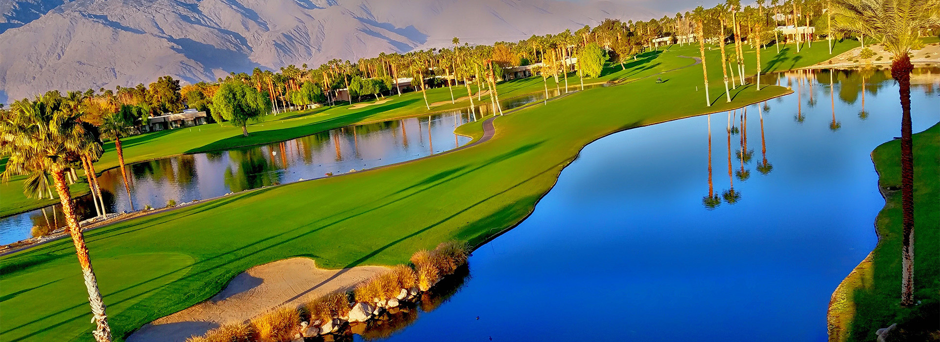 An aerial view of a green fairway, between two water features and a small sand trap. Mountains can be seen in the distance.